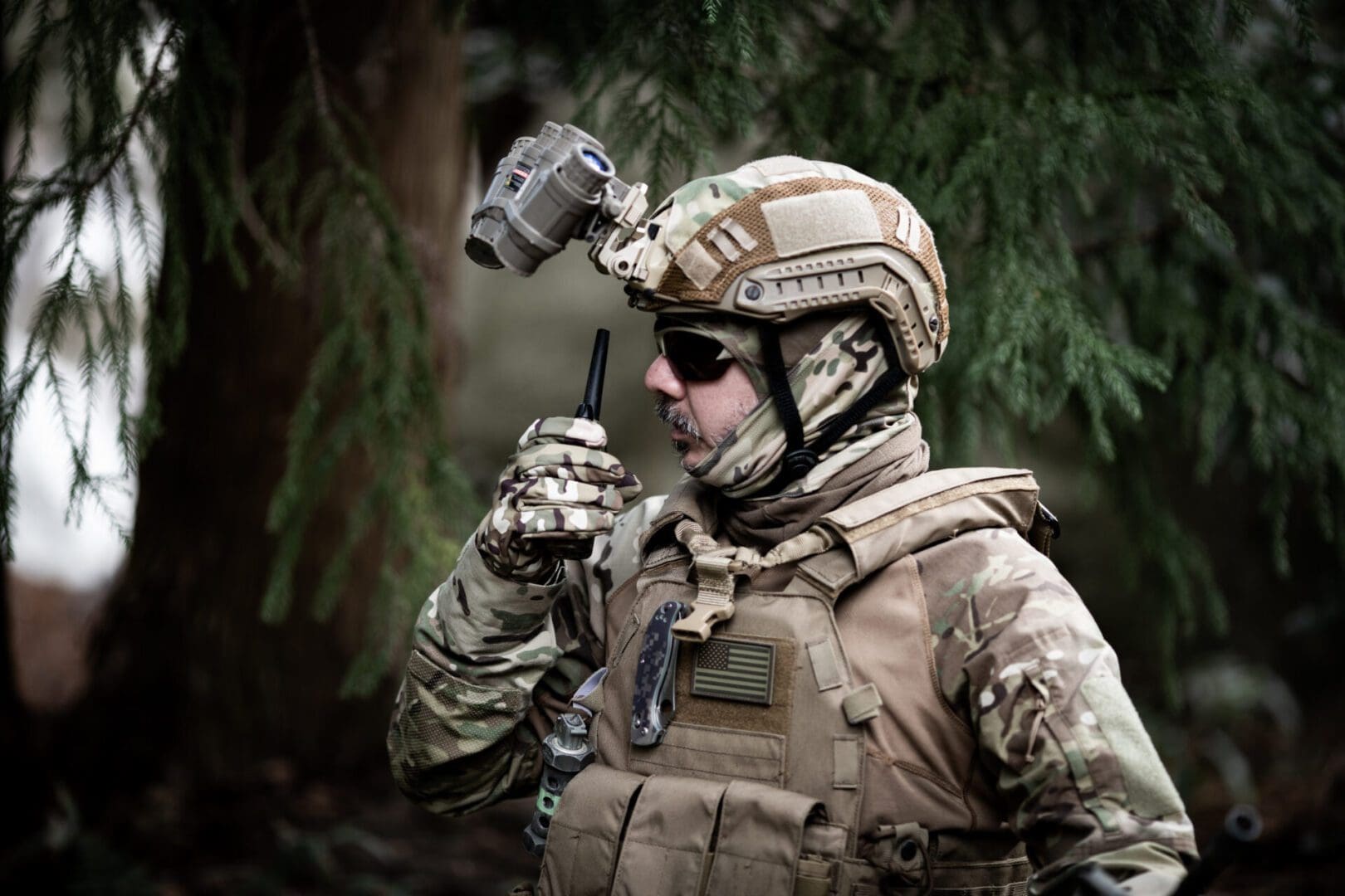 A tactical Military special forces member talking on a walkie talkie in a forest.