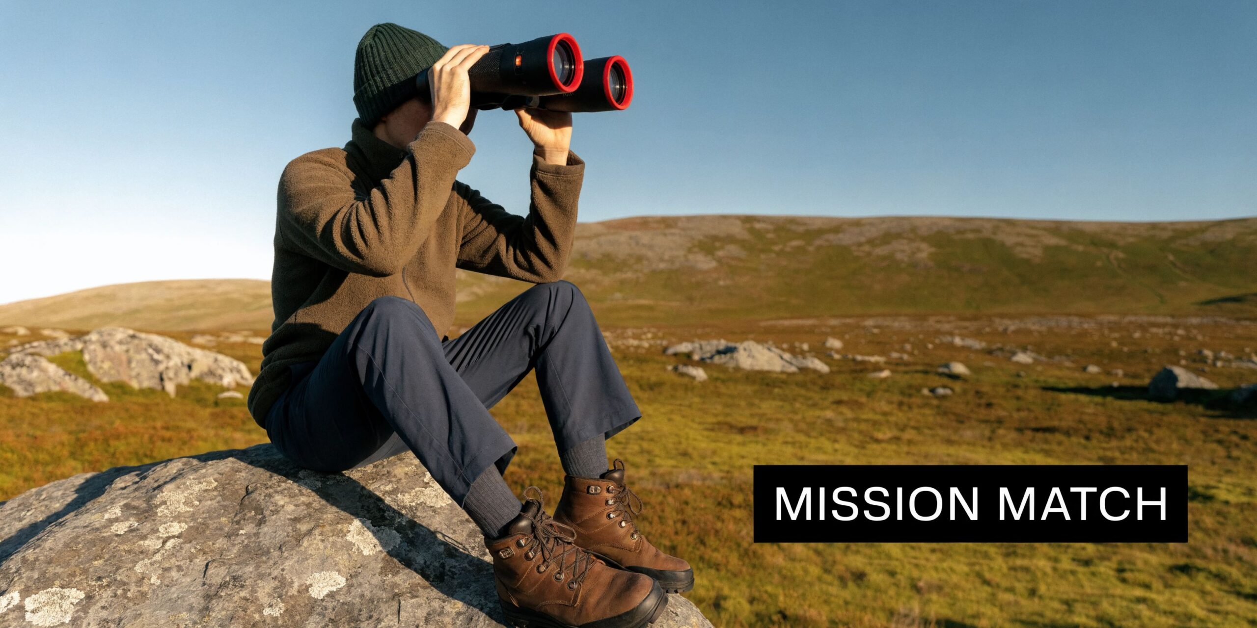 A person sitting on a rock in a landscape, using thermal imaging binoculars to scan the terrain.