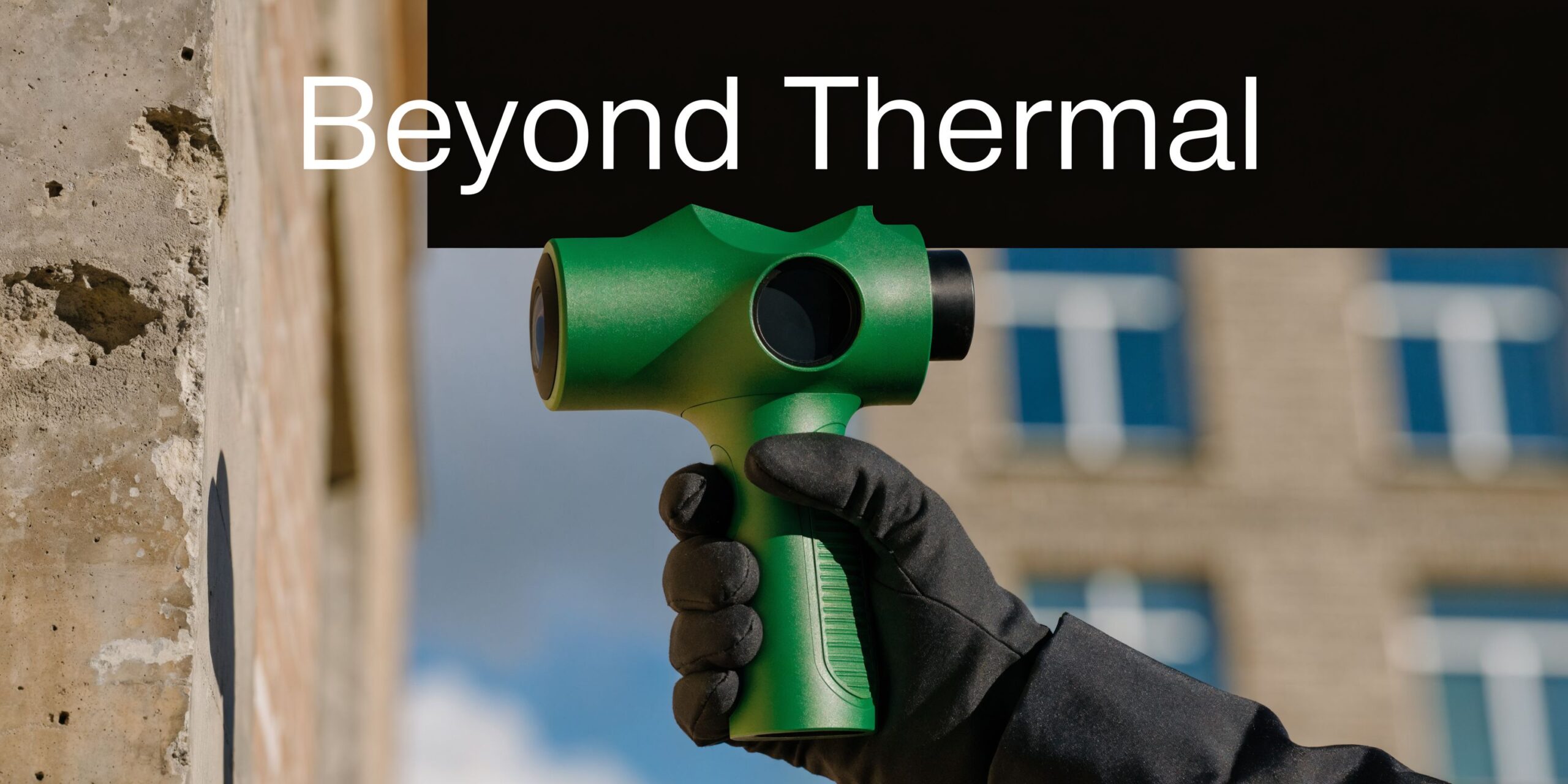A hand wearing a black glove holds a green professional thermal imaging camera against a building wall.