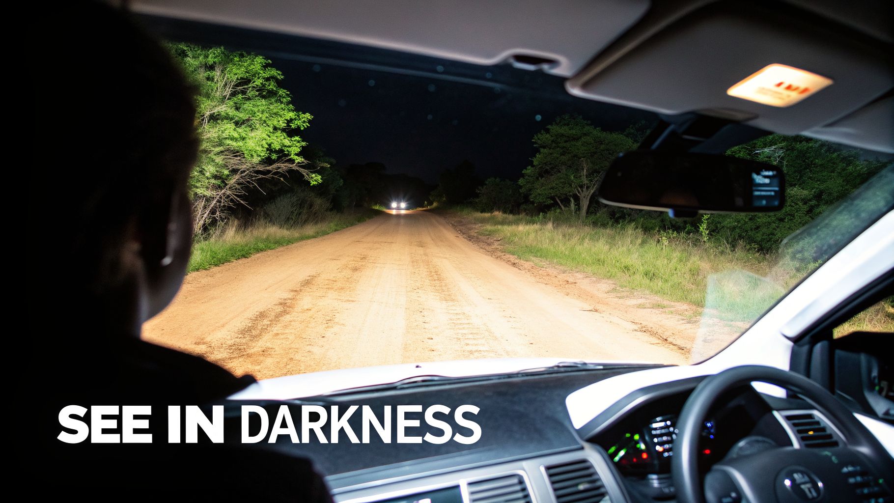 Inside a car at night, bright headlights illuminate a dirt road with another vehicle approaching.