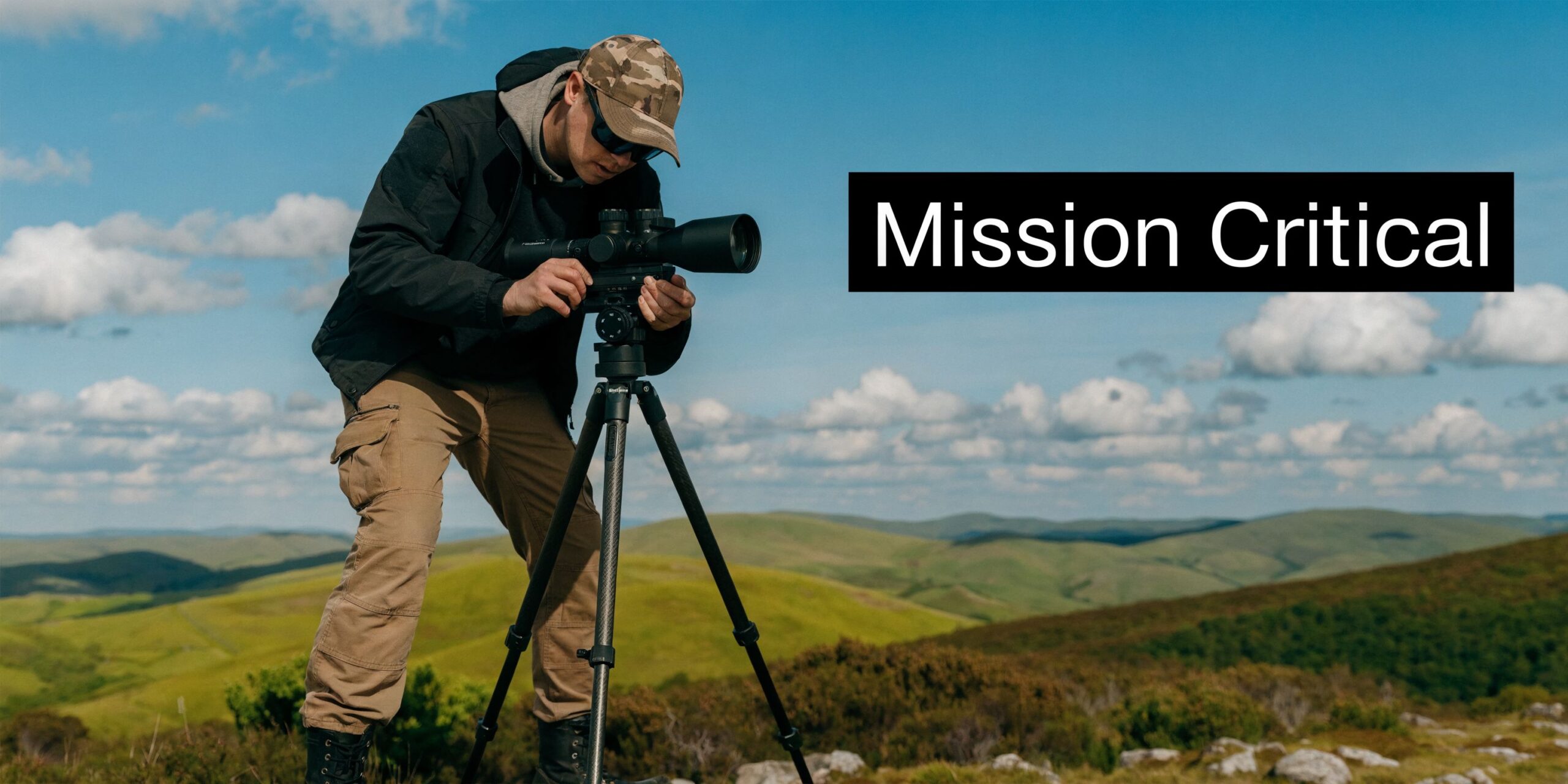 A man in tactical gear adjusting a spotting scope on a tripod overlooking a grassy mountain landscape.
