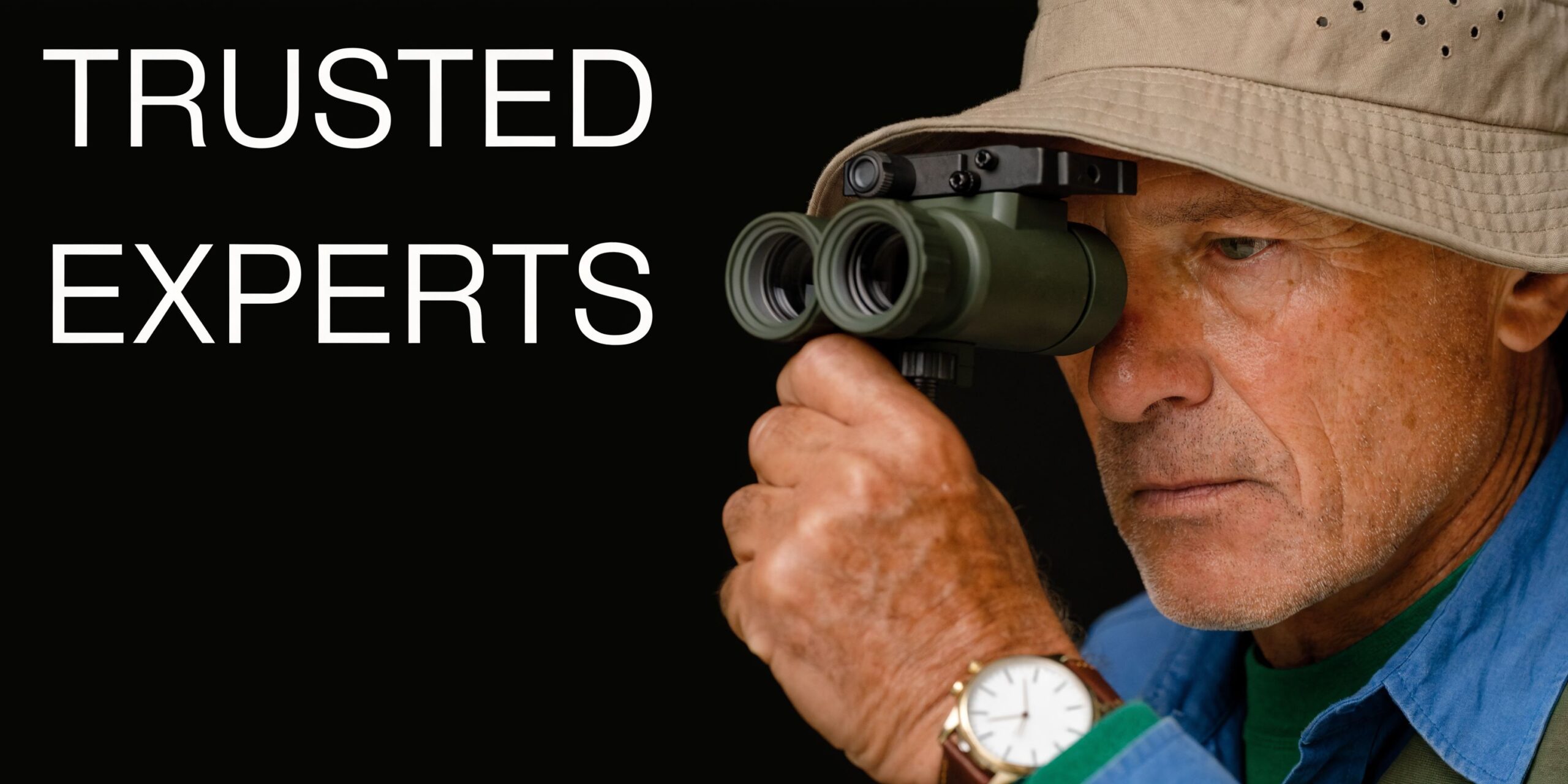 A mature man wearing a hat looks through specialized green night vision binoculars against a black background.